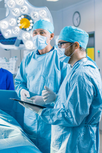 Vertical Shot of a Professional Surgeon and Assistant Talk and Use Digital Tablet Computer During Surgery. They Work in the Modern Hospital Operating Room. - Fotó, kép