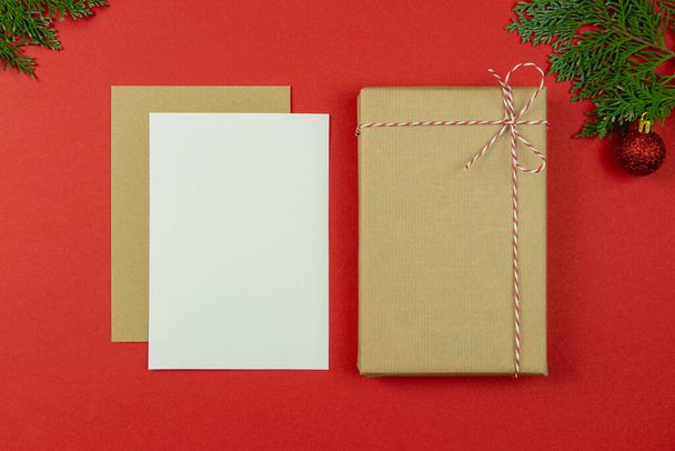 Christmas letter concept. Blank paper card, craft paper envelope, vintage gift box, rope, thuja branches on red background.Holiday Flat lay. Top view. Copy space. Mockup.  - Photo, Image