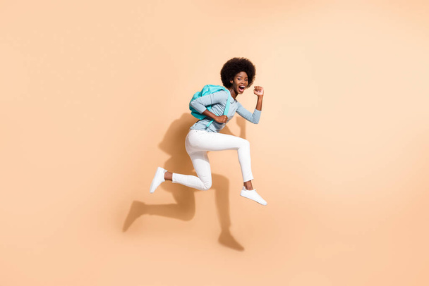 Photo portrait of brunette african american girl running jumping up wearing casual clothes cyan bag isolated on pastel beige colored background - Photo, image