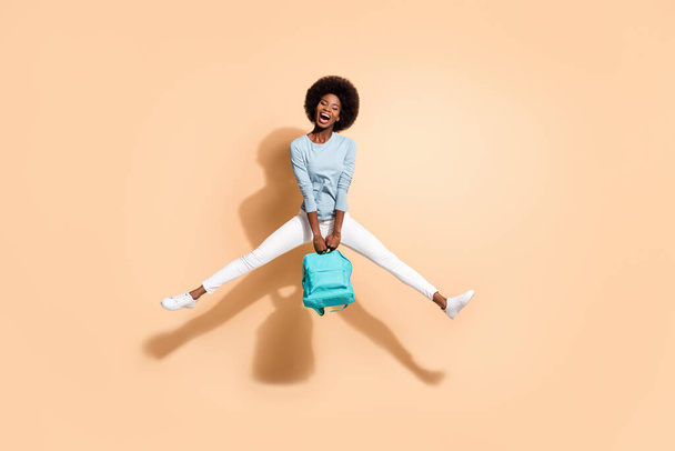 Photo portrait of african american girl holding blue rucksack jumping up with open mouth spread legs isolated on pastel beige colored background - Fotografie, Obrázek