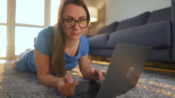 眼鏡をかけた女性が床に横になっていて、クレジットカードやノートパソコンを使ってオンラインで購入しています。オンラインショッピング、ライフスタイル技術 - 映像、動画