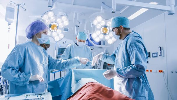 Shot of Diverse Team of Professional Surgeons Performing Invasive Surgery on a Patient in the Hospital Operating Room. Nurse Hands Out Instruments to surgeon, Anesthesiologist Monitors Vitals. Modern - Фото, изображение