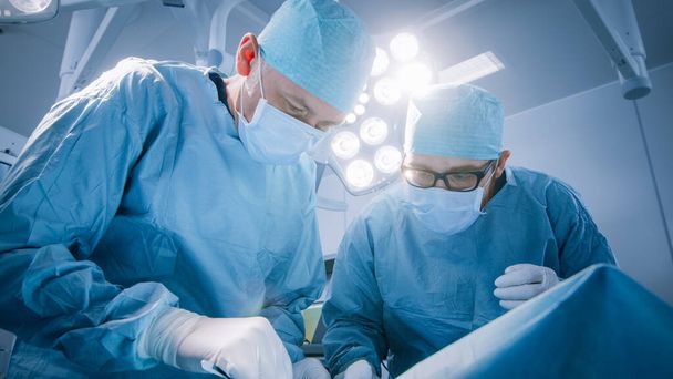 Low Angle Shot In Operating Room of Two Surgeons During the Surgery Procedure Bending Over Patient with Instruments. Professional Doctors in Modern Hospital - Foto, imagen