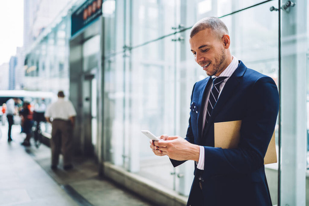 Content stylish business manager in elegant suit with envelop surfing mobile phone near glass wall in light glass business hall - Foto, afbeelding