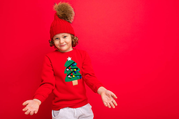 Kırmızı bir Noel süveteri giymiş havalı bir genç kırmızı bir duvarın arka planında, sıcak bir şapkayla ve Noel ağacı olan bir süveterle oynaşıyor. - Fotoğraf, Görsel