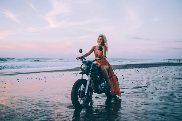 Happy attractive young female with long hair in orange dress sitting on motorcycle and smiling on beach at dusk - Foto, imagen