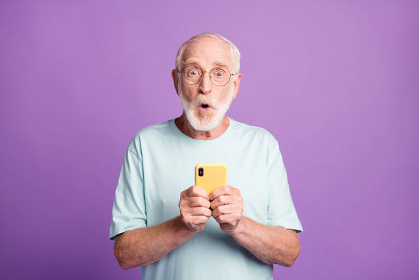 Photo portrait of shocked old man holding phone in two hands isolated on vivid violet colored background - Foto, Bild