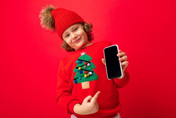 a young man with a smartphone in his hands against the background of a red wall, in a knitted hat and a sweater with a Christmas tree. - Photo, Image