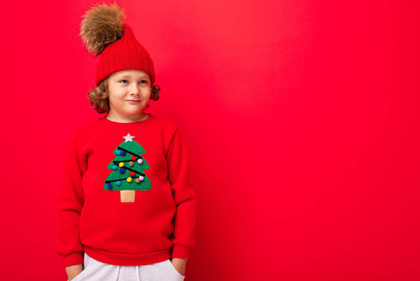 netter blonder Junge mit warmem Hut und Weihnachtspullover auf rotem Hintergrund mit einem Lächeln im Gesicht - Foto, Bild