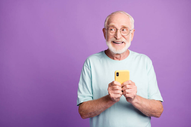 Retrato fotográfico del anciano sosteniendo el teléfono en dos manos mirando el espacio en blanco aislado sobre un fondo de color púrpura vivo - Foto, imagen