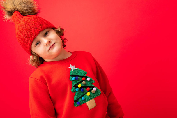cool blond kind in warme hoed en trui met kerstboom op rode achtergrond rommelen rond, kerst concept - Foto, afbeelding