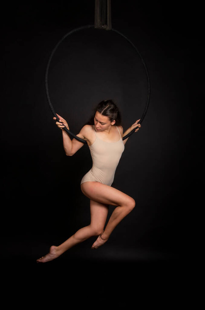 A young woman with long hair is engaged in aerial acrobatics in a beige bodysuit, performs exercises in the air ring . Studio shooting on a dark background. - Foto, Bild