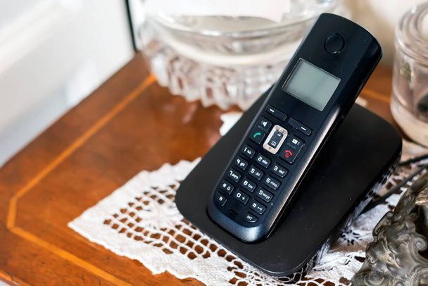 musta muovinen langaton puhelin, joka latautuu kehtoonsa klassisesti sisustetussa kodissa, jossa on antiikkia ja valkoista virkkaa. Vanhentunut tekniikka - Valokuva, kuva