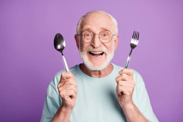 Photo of positive smiling grandfather hold cutlery wear light blue t-shirt isolated over violet color background - 写真・画像