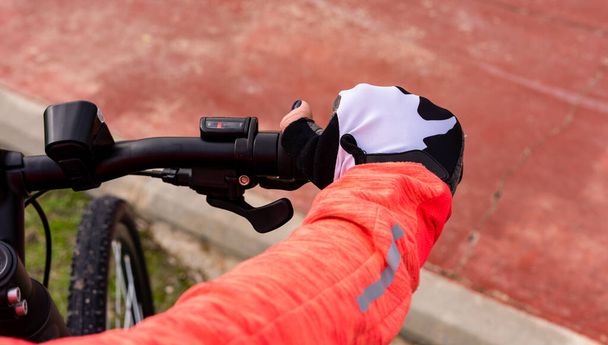 Ženská ruka v oranžové mikině a bezprstých cyklistických rukavicích spočívá na řídítkách bicyklu. - Fotografie, Obrázek