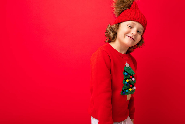 cute blond boy in warm hat and christmas sweater on red background with smile on his face - Zdjęcie, obraz