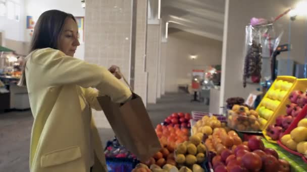 Femme en manteau jaune met une pomme rouge fraîche dans un sac en papier au marché - Séquence, vidéo