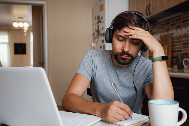 Εστιασμένος νεαρός άνδρας φορούν ακουστικά μελετήσει σε απευθείας σύνδεση βλέποντας webinar podcast σε φορητό υπολογιστή ακούγοντας την εκμάθηση διάσκεψη πορεία καλώντας να κάνει σημειώσεις κάθονται στο γραφείο εργασίας, μαθαίνοντας έννοια - Φωτογραφία, εικόνα