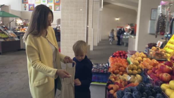 Un niño pequeño con su madre en el mercado. Chico pone melocotones en una bolsa de cuerdas - Imágenes, Vídeo
