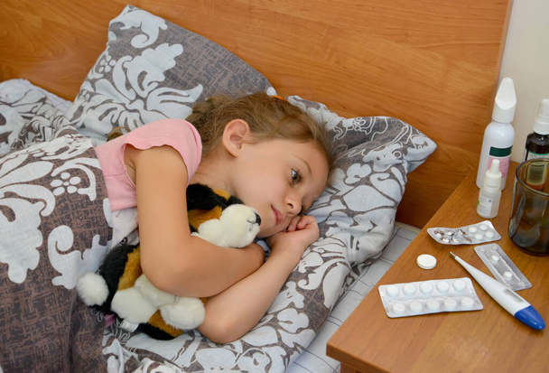 A sad sick girl lies in bed with a soft toy in her hands and looks at the medicine - Foto, immagini