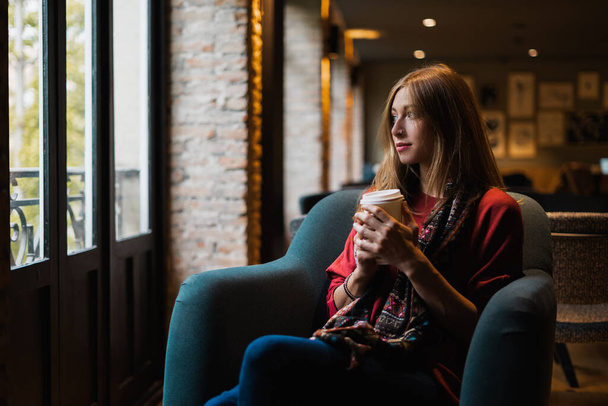 Belle femme blonde prenant un café assis sur un canapé dans une cafétéria regardant par la fenêtre. - Photo, image