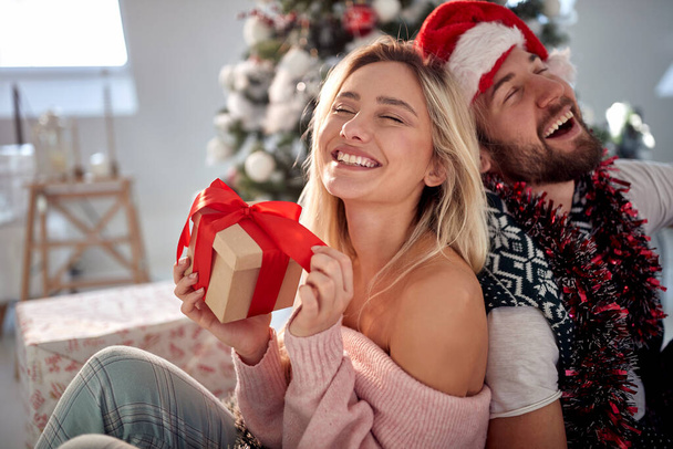 gelukkig jong paar leunen rug aan rug, lachen, het vasthouden van cadeautjes in de voorkant van kerstboom - Foto, afbeelding