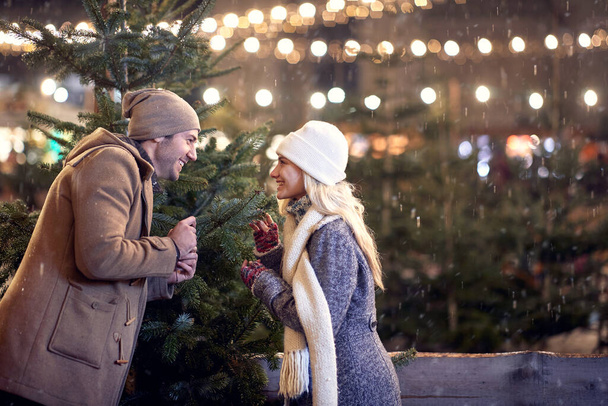 A young cheerful couple looking for a perfect christmas tree in a magical night on a snowy weather in the city. Christmas tree, love, relationship, Xmas, snow - Zdjęcie, obraz