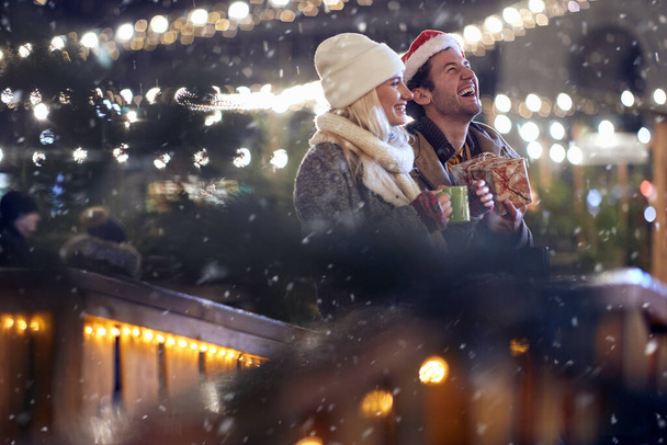 A young couple in love enjoying snowfall in a magical night at christmas festival in the city. Festival, love, relationship, Xmas, snow - Foto, afbeelding