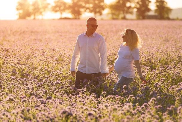 Beautiful pregnant mother and her husband walk together hand in hand outdoor, in the middle of a flowery field at sunset time. - Fotografie, Obrázek