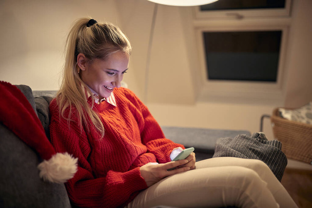 portrait of young caucasian blonde sitting on sofa using her cell phones on christmas, smiling - Fotoğraf, Görsel