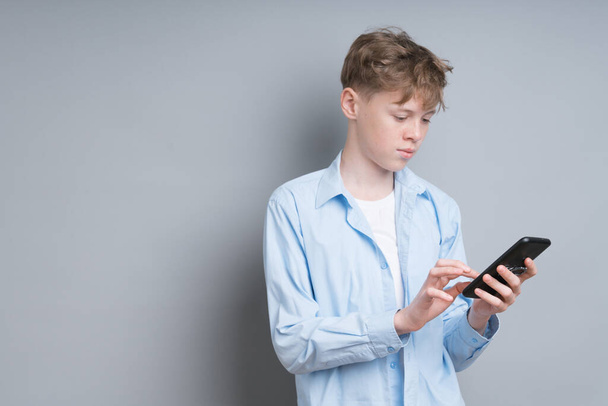 A teenager boy in a blue shirt and a white T-shirt plays the phone on a gray background. Troubled teenager, social issues, gamer, phone addiction - Foto, imagen