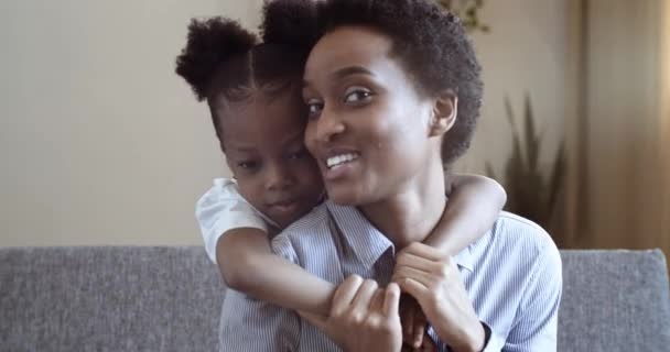 Portret van twee mensen Afrikaanse moeder en schattige kleine kleuter dochter praten lachen samen knuffelen zitten in de woonkamer bank. kind meisje knuffels mam achter haar rug liefdevol familie concept - Video