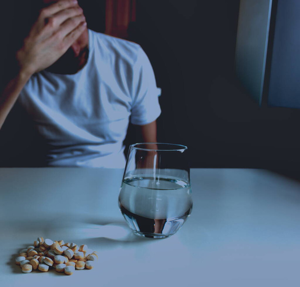 Aşırı dozda hapla intihar etmeyi düşünen depresyonlu bir adam, beyaz masada kasıtlı olarak karartılmış arka planda oturuyor. - Fotoğraf, Görsel