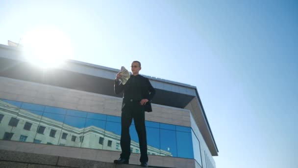 Business man manager worker wears suit, stands outdoor near modern blue glass building, holds dollars in his hands, waves fan of money to his face, confident businessman feels success, have salary - Footage, Video