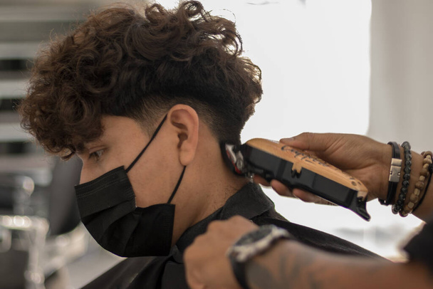 Bir kuaförde maske takmış genç bir kuaförle seans. Salgın zamanlarında maskeli bir çocuğun saçını kesiyor. - Fotoğraf, Görsel