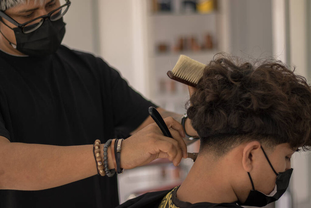 Séance dans un salon de coiffure avec un jeune coiffeur avec un masque coupant les cheveux d'un garçon avec un masque en période de pandémie - Photo, image