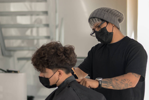Séance dans un salon de coiffure avec un jeune coiffeur avec un masque coupant les cheveux d'un garçon avec un masque en période de pandémie - Photo, image