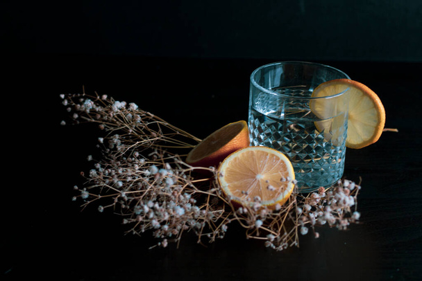 Alcohol drink (gin and tonic cocktail) garnished with lemon fruit and flower isolated on black background. Iced cocktail drink with lemon and herbs. - 写真・画像