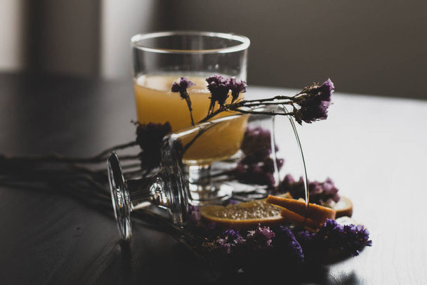 パルプとスライスされた果物を含む健康的なオレンジジュースのガラスは、紫色の花の装飾と日光と木製の背景に隔離されます。オレンジジュースとテーブルの上の新鮮な果物. - 写真・画像