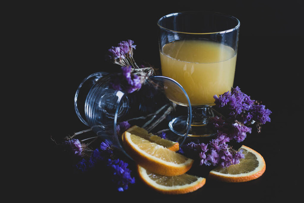 果肉とスライスされた果物の健康的なオレンジジュースのガラスは、紫色の花で黒の背景に隔離されます。オレンジジュースとテーブルの上の新鮮な果物. - 写真・画像