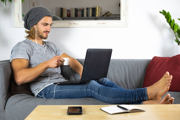 Przystojny młody biznes biały człowiek ze słuchawkami pracujący w domu za pomocą laptopa i pijący filiżankę herbaty - Zdjęcie, obraz