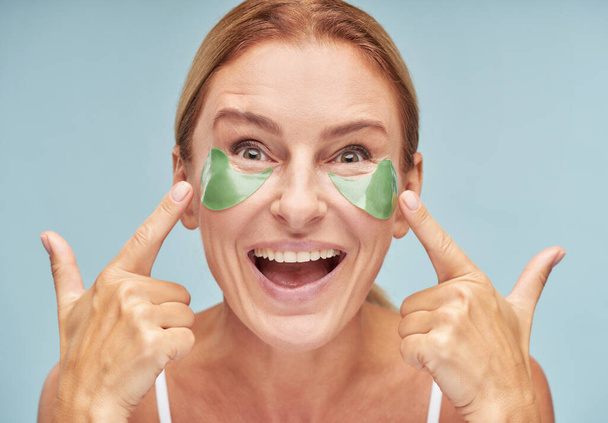 Happy mature lady moisturizing skin around her skin while using patches - Photo, Image