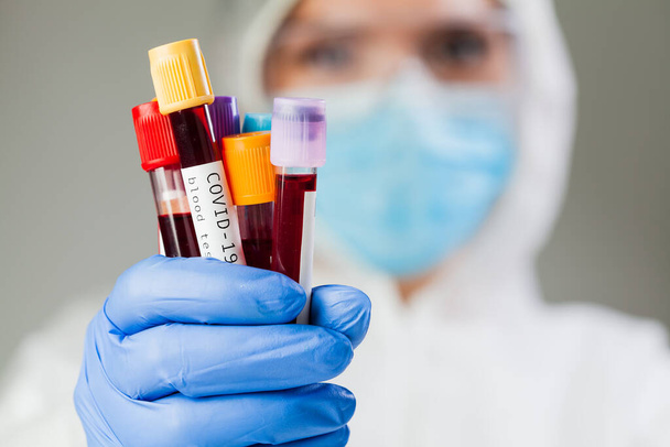 Лаборант или медицинский технолог в защитном костюме и маске, в котором содержатся вакуутеры пробирки, образец крови пациента с коронавирусом, визуализация вируса COVID-19 ПЦР анализа ДНК - Фото, изображение