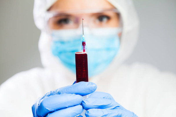 Lékař nebo laboratorní vědec v osobních ochranných pomůckách držící stříkačku plnou krevní plazmy, se zaměřením na jednu kapku kapající z hrotu jehly, léčbu a výzkum očkovací látky - Fotografie, Obrázek
