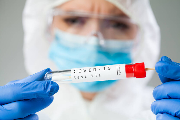 Медицинский технолог, держащий набор для сбора мазков COVID-19, маску и перчатки белого защитного костюма, пробирку для взятия образца образца пациента ОП, иллюстрацию процесса тестирования ДНК ПЦР - Фото, изображение
