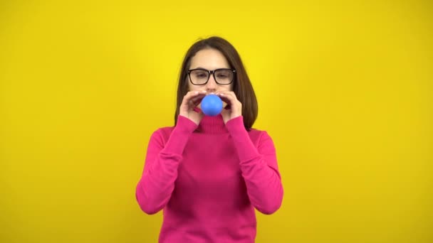 Młoda kobieta nadmuchuje niebieski balon ustami na żółtym tle. Dziewczyna w różowym golfie i okularach. - Materiał filmowy, wideo