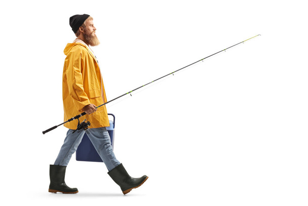 Full length profile shot of a bearded fisherman walking and carrying a fridge and a fishing rod isolated on white background - Fotó, kép