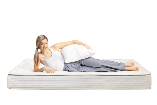 Young woman lying on a bed mattress in pajamas and holding a pillow isolated on white background - 写真・画像