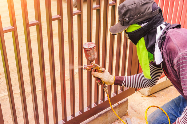 Mannelijke werknemers gebruiken een verfspuit bij een stalen deur. Werknemers moeten spuitverf beschermen tegen inademing door het gebruik van een doek om de neus te bedekken. - Foto, afbeelding
