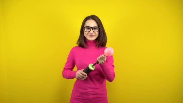 Mladá žena nafoukne růžový balón čerpadlem a to praskne na žlutém pozadí. Dívka v růžovém roláku a brýle. - Záběry, video